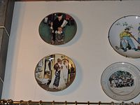 Photo showing four decorative plates displayed on a wall depicting scenes such as a man and child reading, and a man mowing a lawn.