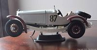 Side profile of white vintage Mercedes-Benz SSKL model car on black stand showing details of wheels, number 87, and red fenders.