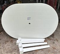 Large white plastic oval outdoor table top with four white plastic legs detached, lying on ground.