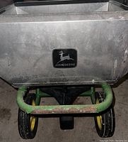 Side view of the John Deere green fertilizer/grass seed spreader showing rust on the green frame and black John Deere logo on the silver hopper.