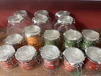 Photo showing 16 clear glass storage jars with clamp lids arranged on a wooden surface, some filled with various contents. Jars vary in size and have labels showing volume and LeJar branding.