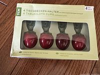 Set of 4 apple-shaped red plastic tablecloth clips in original box, showing hand-painted design and weatherproof plastic material