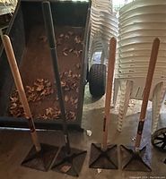 Front view of four tampers with square metal heads and wooden and black handles leaning against stacked chairs and a wheelbarrow, showing size and condition.