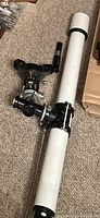 White and black metal refractor telescope tube with mounting assembly, adjustment knobs, and angled eyepiece.