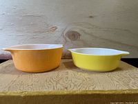 Two Pyrex CorningWare bowls side by side on a wooden surface. One bowl yellow, one orange, both with pouring spouts and white interiors.