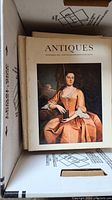 Box containing multiple issues of Antiques magazines, showing one front magazine cover with painting of a woman in period dress