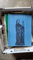 Photo of blue cover Antiques magazine showing a statue of a woman holding a child, along with other magazines in a box.