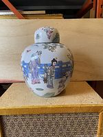 Front view of the porcelain jar showing hand-painted story figures and architectural elements on rounded body with lid on top.