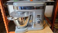 Cuisinart silver stand mixer with attached stainless steel mixing bowl placed on a shelf.