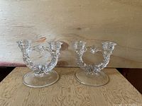 Pair of clear glass double arm candle holders on wooden surface with wood background.