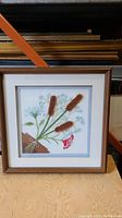 Framed woven floral textile artwork showing full piece with light brown wooden frame and white matting on table in storage.