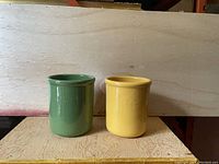 Photo showing two cylindrical Emile Henry utensil holders in yellow and green colors, side by side on a wooden surface.