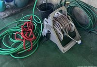 Four garden hoses with one mounted on beige plastic wind-up reel; other hoses coiled around the reel, on the ground, and nearby wall