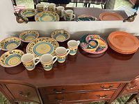 Photo showing four Royal Norfolk dinner plates, bowls, mugs, five rust colored dinner plates stacked, and multicolor divided serving dish on wooden sideboard with mirror background.