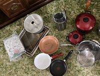 Photo showing assorted cookware including metal steamer pots, colanders, sauté pans, mini muffin pan, cookie sheets, and cooling rack arranged on carpet.