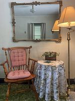 View of traditional maple wooden chair with tufted pink cushions, round table with floral tablecloth, gilt edged mirror, and brass floor lamp