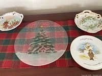 Holiday serveware displayed on a red and green plaid cloth, including a clear glass platter with an embossed Christmas tree, and handpainted candy dishes with poinsettia and holly designs, plus a Holly Hobbie plate with a holiday scene.