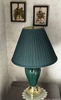 Green ceramic ginger jar lamp with brass base and dark green pleated fabric shade, shown on a small table