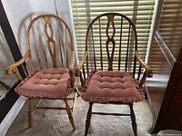 Pair of maple Windsor style chairs with curved spindle backs and armrests, rose-colored tufted cushions, placed indoors near window with blinds.