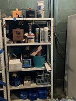 Seven shelves filled with assorted painting and home repair supplies including paint cans, boxes, and tools, arranged in a basement area with cinder block walls