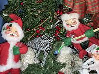 Photo showing two Annalee Santa dolls, one holding skis and ski poles, set among green pine garland and festive berries, silver wireframe decorations visible.