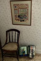 Photo showing the wooden upholstered chair, needlepoint artwork on the wall, and two metal trashcans with distinct designs.