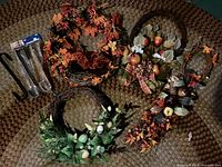Image of eleven assorted decorative wreaths and three black metal over-the-door wreath holders arranged on a braided rug. Wreaths include autumn leaves, floral, twig, and seasonal themes mostly handmade.