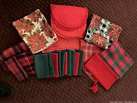 Holiday-themed tablecloths, napkins and runners arranged on carpet showing various red, green, plaid, poinsettia, and floral fabrics.