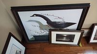 Photo showing two framed prints leaning against a wall on a wood floor. One larger print depicts a black bird sculpture with red-berried branches, the other is a smaller black and white photo print.