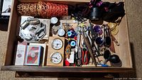 Top view of drawer showing multiple items including chess clock, playing cards, tools, pins, sunglasses, headlamp, glass decoration
