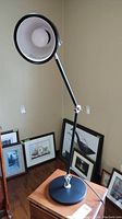Black metal adjustable desk lamp standing on a wooden surface with framed pictures in the background. Round base, adjustable arm with three silver joints, and conical black shade with white inner lining.