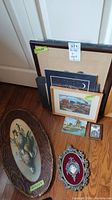 View of assorted framed items leaning against wall and floor, including large oval decorative tile with pheasants, various framed prints, small framed photograph, and a red/silver ornamental piece.