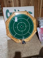 Crokinole game board shown with green playing surface and scoring rings on wooden octagonal frame, game pieces in front in plastic bag.