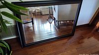 Framed mirror shown leaning against wall on hardwood floor with visible indoor plants and furniture reflections.