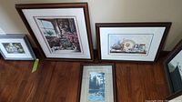 Photo showing three framed prints leaning against a wall on wooden floor. The largest is Laura Berry's "Frills and Flowers" featuring flowers by a window, numbered 617/750 and signed. Next to it is a framed print of decorative plates and shells titled "The Garden Shed", numbered 361/750 and signed. A smaller nature scene print is visible in front.