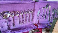 Silver plated spoons, forks, and serving utensils secured in a purple fabric roll organizer, showing the layout and item types present.