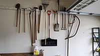Full view of multiple garden tools hanging on wall hooks showing pitchfork, shovels, rakes, snow shovel, saws, and pruner