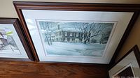 Framed print showing snowy house under twilight sky with signature and numbering visible.