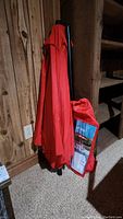 Photo of folded red umbrella leaning against wood wall, shows umbrella closed and bag beside it.