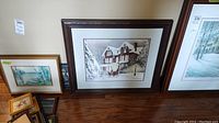 Framed print depicting winter house scene with people, alongside other framed artworks, showing overall size and setting.