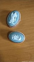 Two oval Wedgwood Jasperware brooches on wooden surface, showing front relief designs and gold-tone metal frames.