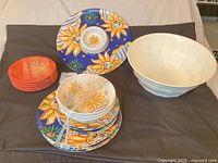 Group view of sunflower pattern melamine platter, plates, bowls, stack of orange plates, and white salad bowl