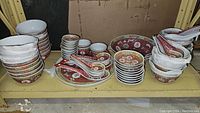 Stacks of plain white bowls, red and gold rimmed bowls, small plates and soup spoons on a shelving unit