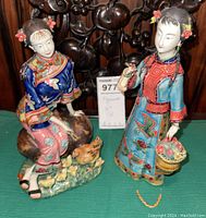Photo showing both Asian geisha figurines on green surface, one standing holding a basket and bird, the other seated with birds at feet.