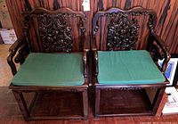 Pair of vintage Asian rosewood armchairs with green cushions shown side by side against a wood paneled wall. Carved backrests with intricate floral and emblematic detailing. Solid armrests and rectangular legs.