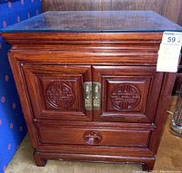 Front view of one vintage Chinese mahogany/rosewood side table showing cabinet doors with circular carvings, brass hardware, drawer and glass top.