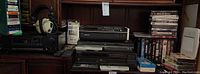 Wide shot of DVD player, CD player, taped VHS and DVDs, headset on shelf with remotes