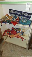 Shelf with multiple pennant flags and a 'Bangka Bersatu' blue felt banner, mixed sizes and colors.