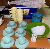 Various kitchenware including cream mugs, blue tea cups with saucers, white bowls, green bowl and Igloo cooler on table with umbrella above