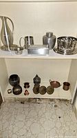 Shelf view showing multiple assorted metal ware items including pitchers, urns, bowls, and boxes with tarnish and wear.